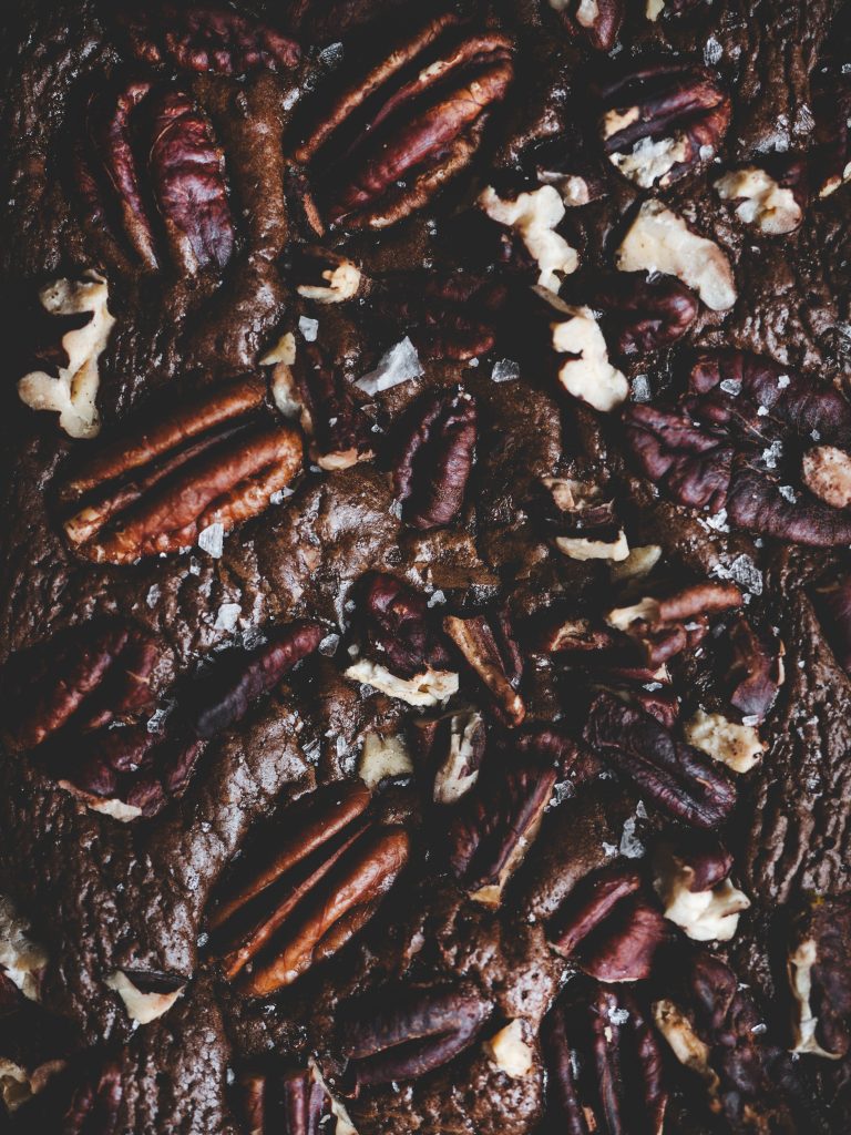 Dettaglio della superficie di un brownie al cioccolato con noci pecan, granelli di sale e pezzi di cioccolato fondente.