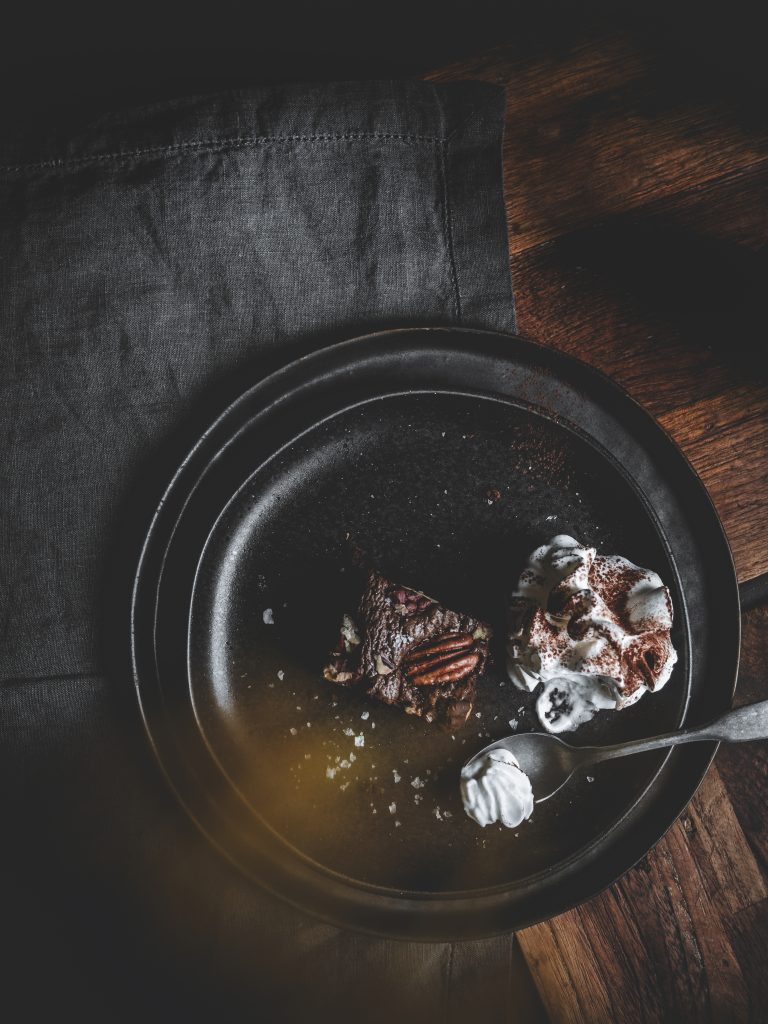 Brownie al cioccolato con noci pecan servito con panna montata su piatto nero e cucchiaio.