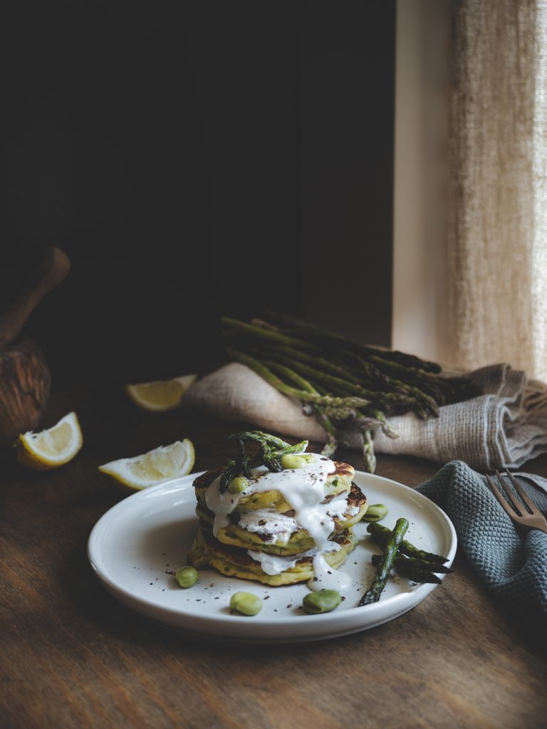 Pancake salati di primavera con fave e asparagi serviti a torre con crema di pecorino e pepe nero su tavolo rustico.