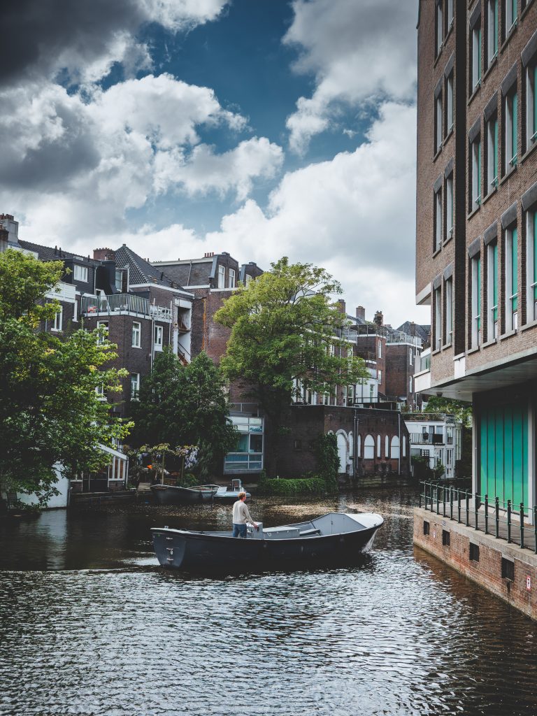 Piccola barca lungo i canali di Amsterdam circondata da edifici storici, alberi e cielo nuvoloso.
