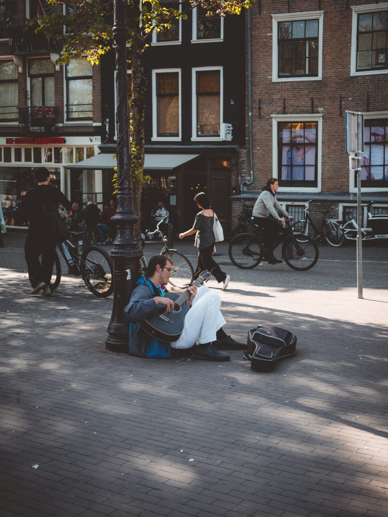 Musicista di strada seduto vicino a un canale di Amsterdam mentre suona la chitarra tra biciclette e luce del pomeriggio.